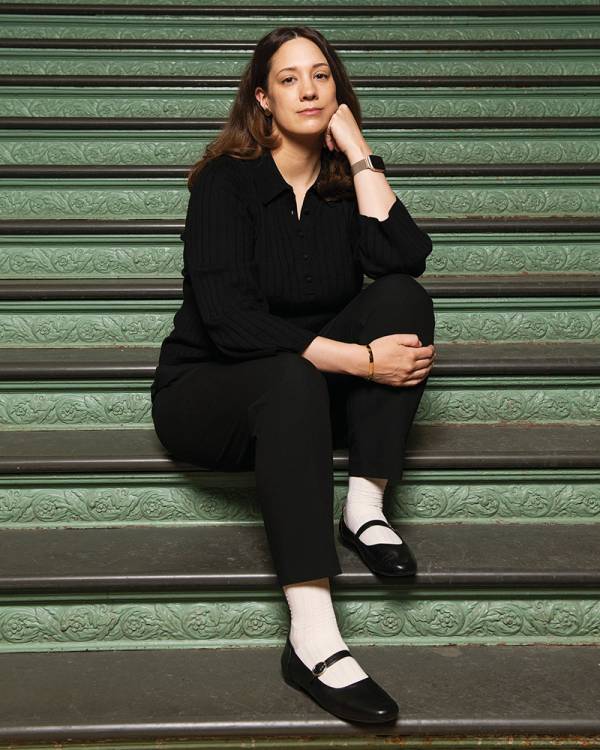 Karina Bold, wearing a black, button-up blouse, black pants and black Mary Jane flats, sitting on a set of stairs with green, leaf-patterned moulding between dark grey steps