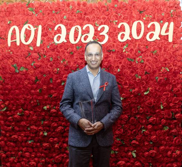 Rajiv Singal stands in front of a photo wall that is covered in red flowers with the words "AOI 2023-2024" in white script at the top of a wall. Singal is holding a clear vase, his award, and smiling