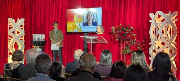 UC Acting Principal Sali Tagliamonte welcomes guests and honourees to the AOI Awards on a stage in front of red backdrop. 