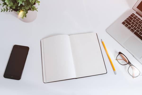 An open notebook on a white desk next to a phone and laptop