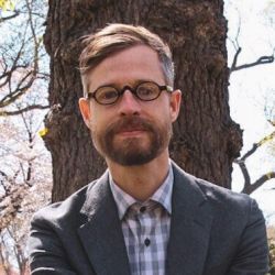 Mark Miller wearing a grey jacket with a white-blue striped shirt, and round glasses.