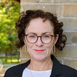 middle-aged caucasian woman with short curly hair wearing white shirt, black blazer, and round dark brown glasses