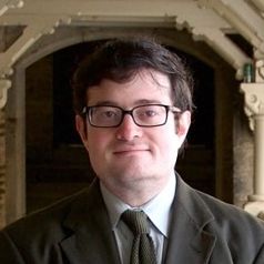 Midshot of Joran Bear, wearing a black suit, and glasses.