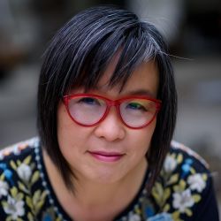 A picture of Larissa Lai smiling. She is wearing specs with red frames and a floral patterned shirt.