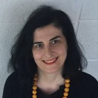 Giancarla Periti headshot, smiling at camera wearing a black sweater, with a yellow beaded necklace on.