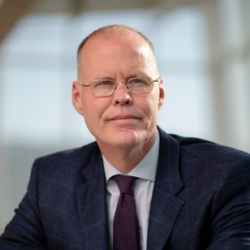 Adalsteinn Brown, wearing a suit and grey glasses, staring of to the side of the camera.