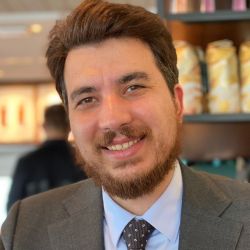 Headshot of Can Mekik, wearing a grey jacket with a blue shirt and grey tie, smiling at the camera.