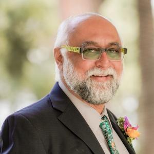 Portrait of Amir Hussain smiling at the camera, wearing a dark grey suit, pale grey shirt and bright blue and green impressionist floral tie with an orange and pink boutonnière in his lapel. H