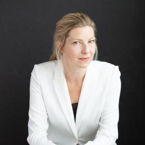 Portrait of Maev Beatty, a woman with blond hair pulled gently back and wearing a natural look in a crisp white blazer with a black shirt barely visible. Her face is turned slightly to the left but she is looking at the camera with a slight smile.