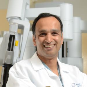 Dr. Singal wears a white lab coat, seated in front of a Da Vinci Surgical System machine. He is smiling at the camera with his arms folded in his lap.
