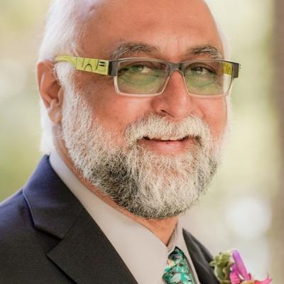 Portrait of Amir Hussain smiling at the camera, wearing a dark grey suit, pale grey shirt and bright blue and green impressionist floral tie with an orange and pink boutonnière in his lapel. H