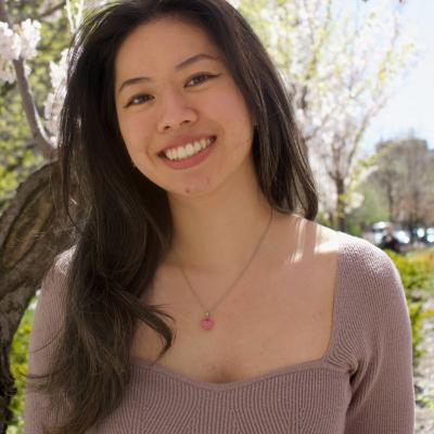 Outdoor portrait of Diana Lam, her head tilted slightly to the right as she smiles at the camera. in a beige rose shirt with her long black hair falling over her right shoulder. The out-of-focus tree behind her appears to be in bloom.