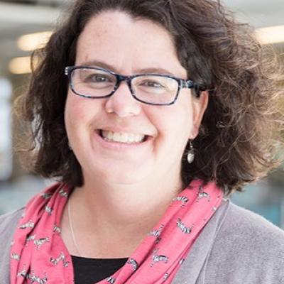 Portrait of Julie Drolet smiling, her face framed by her short wavy brown hair and oval shaped glasses. She is wearing a light pink scarf.