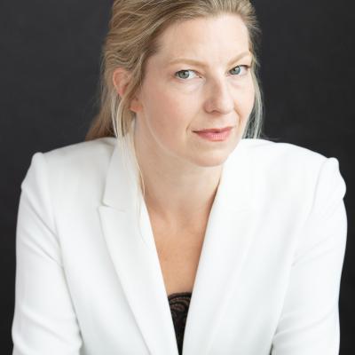 Portrait of Maev Beatty, a woman with blond hair pulled gently back and wearing a natural look in a crisp white blazer with a black shirt barely visible. Her face is turned slightly to the left but she is looking at the camera with a slight smile.