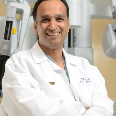 Dr. Singal wears a white lab coat, seated in front of a Da Vinci Surgical System machine. He is smiling at the camera with his arms folded in his lap.